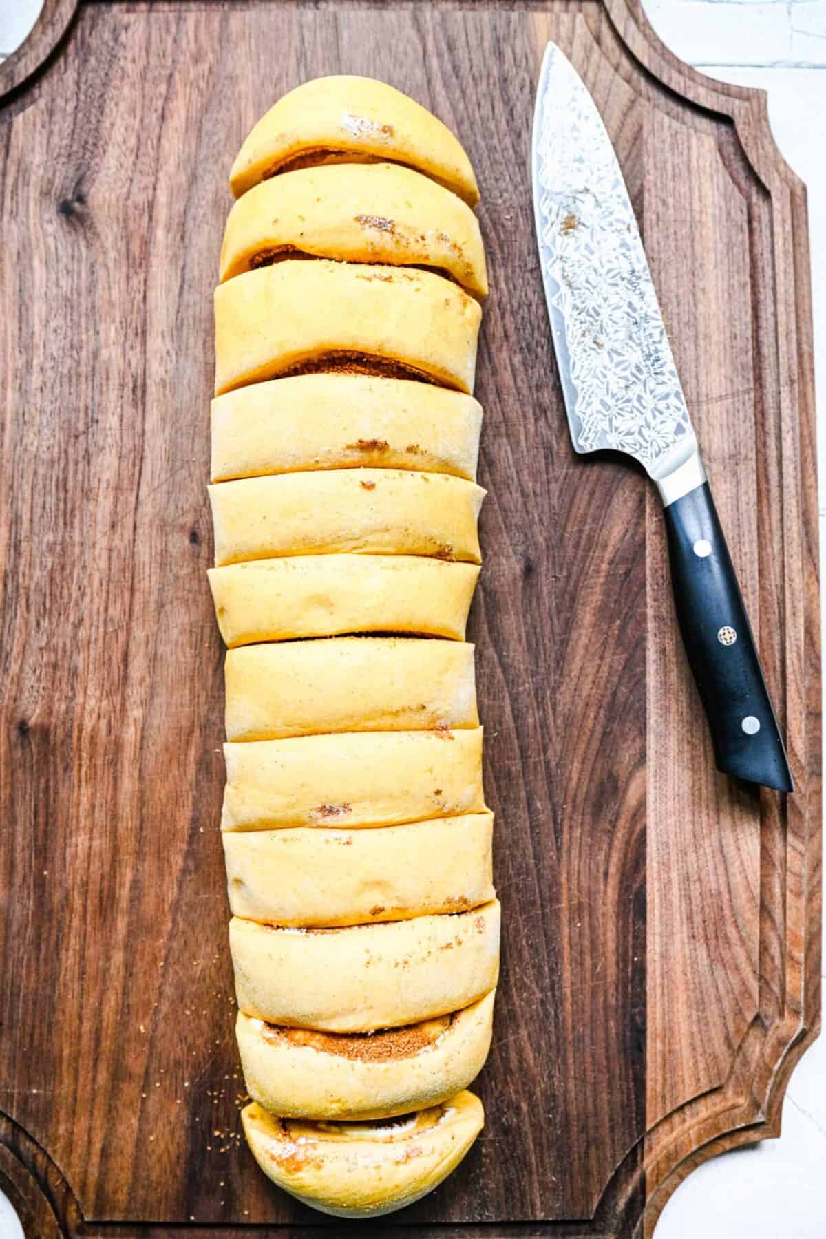 An uncooked log of cinnamon rolls, cut into 12 pieces, on a cutting board next to a chef's knife