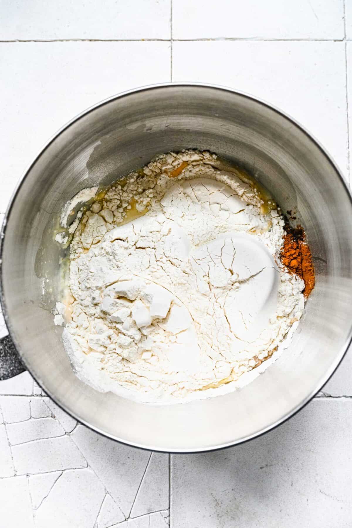 A stand mixer bowl with unmixed ingredients for pumpkin cinnamon roll dough, with flour on top