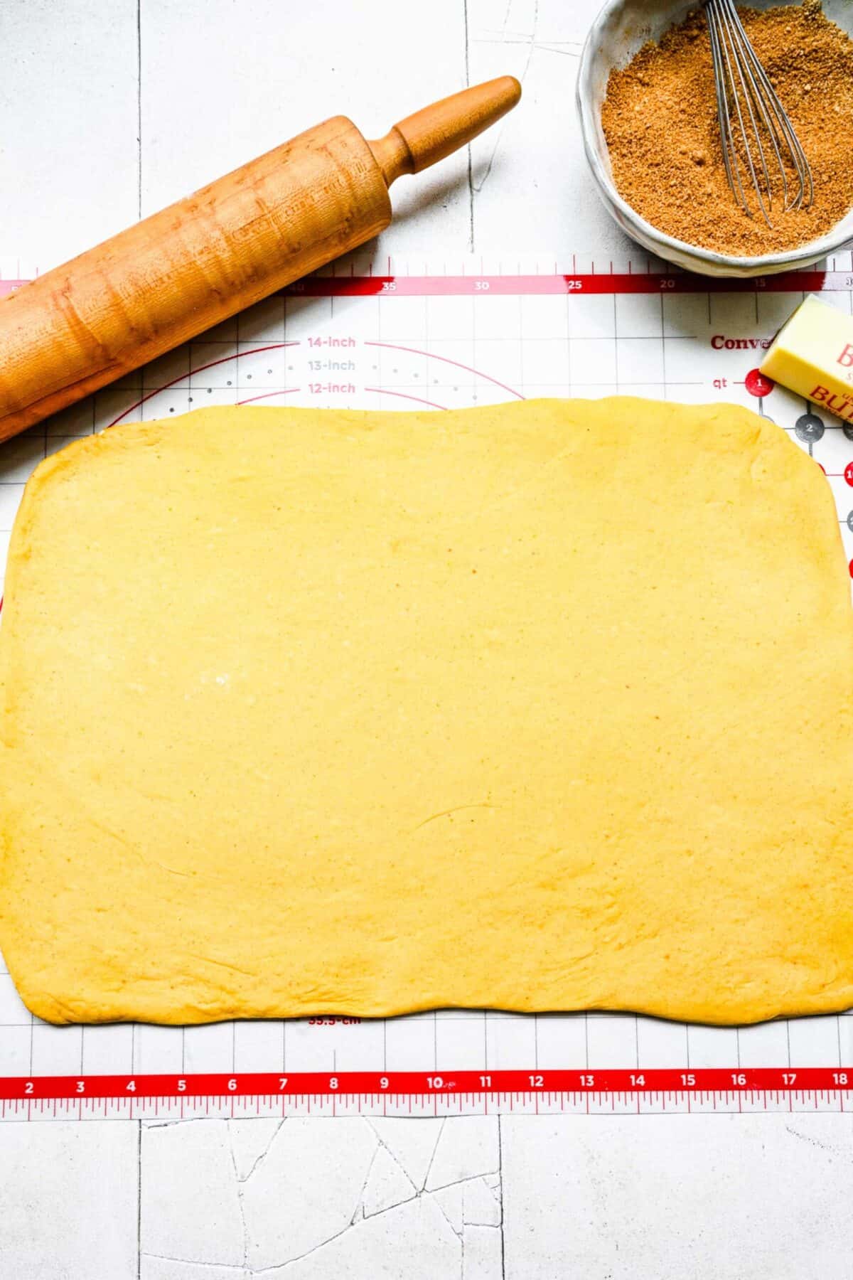 A rectangle of pumpkin cinnamon roll dough on a ruler mat, next to a rolling pin, a stick of butter, and a bowl of spice mixture