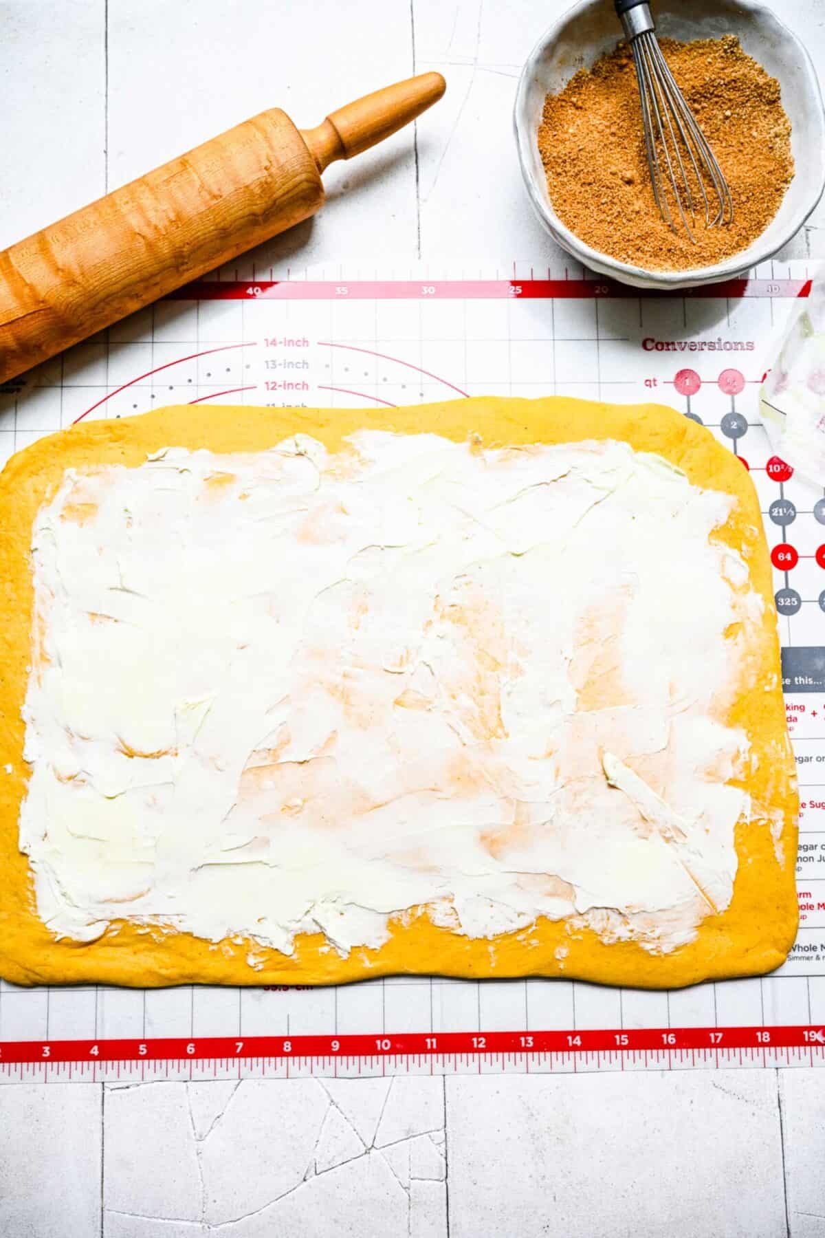 A rectangle of cinnamon roll dough covered in a layer of butter on a ruler mat, next to a rolling pin and a bowl of spice mixture