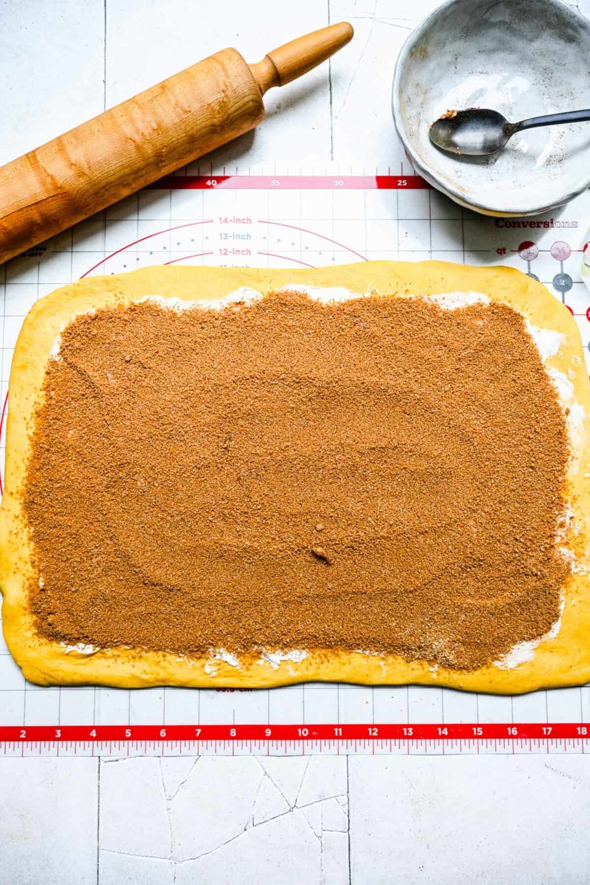 A rectangle of pumpkin cinnamon roll dough covered with a layer of butter and a spice mixture on a ruler mat, next to a rolling pin and an empty bowl with a spoon in it
