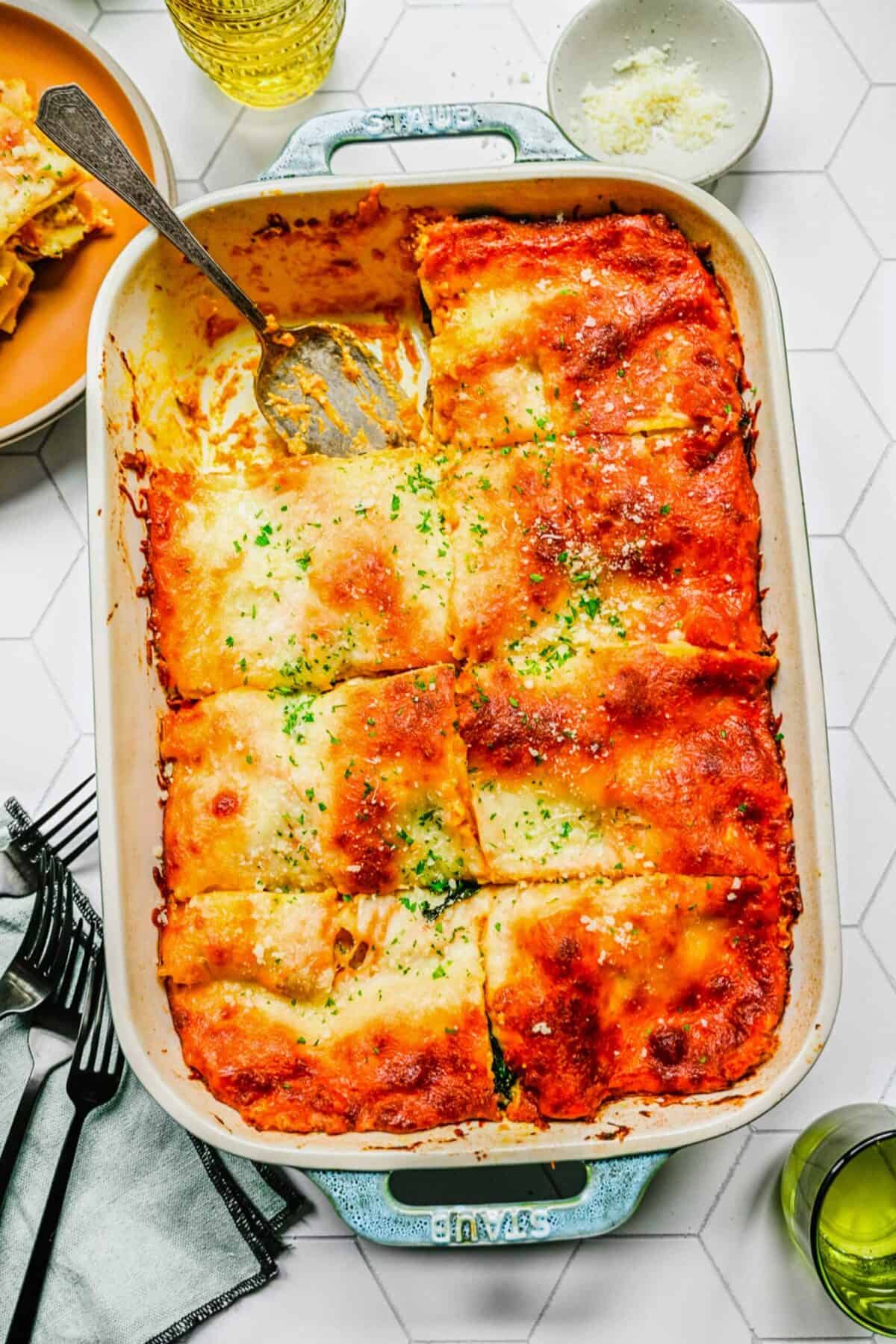 A baking tray with pumpkin lasagna cut into eight slices, with one slice removed and a serving spoon in it