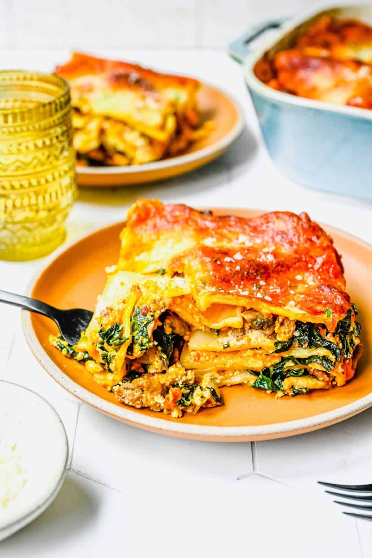 A plate with a slice of pumpkin lasagna, with a fork taking a forkful of it, with another plate of lasagna in the background