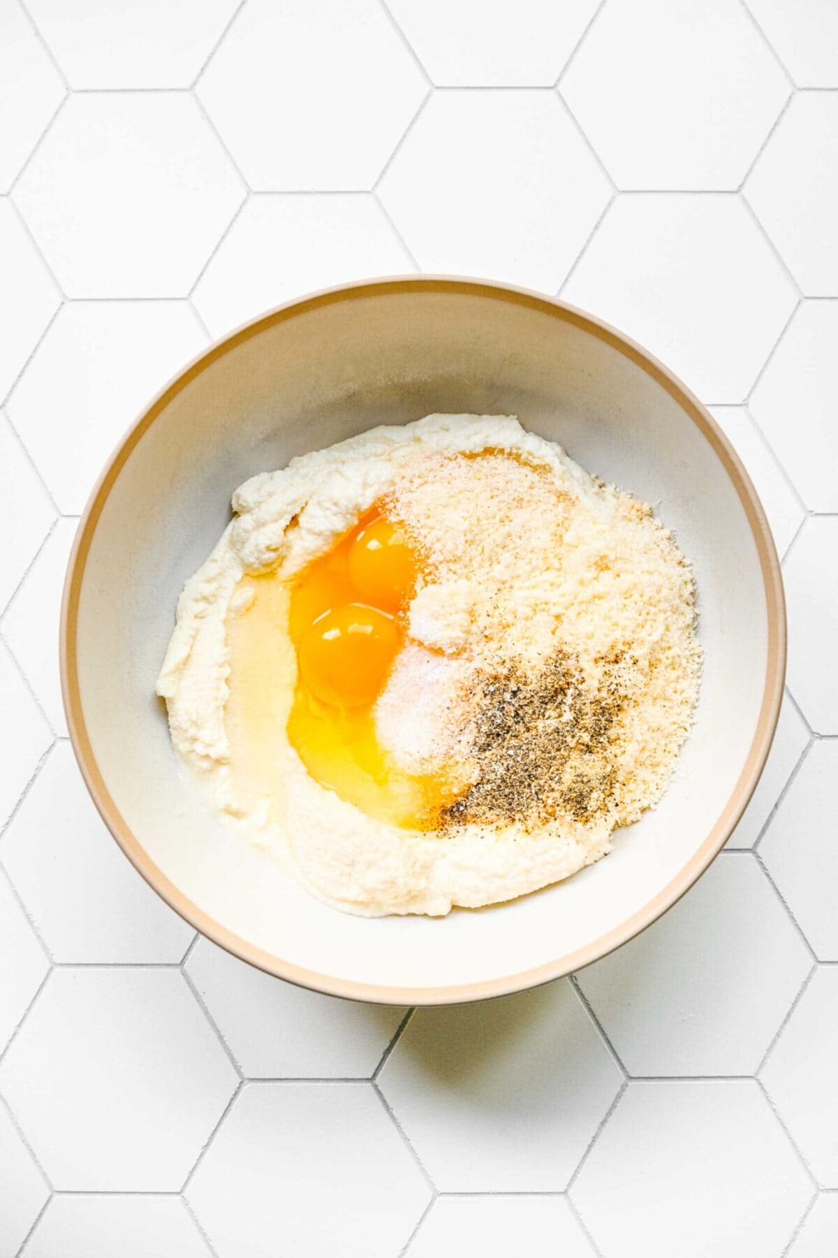 A mixing bowl filled with ricotta, grated parmesan, eggs, salt, pepper, and garlic powder, unmixed