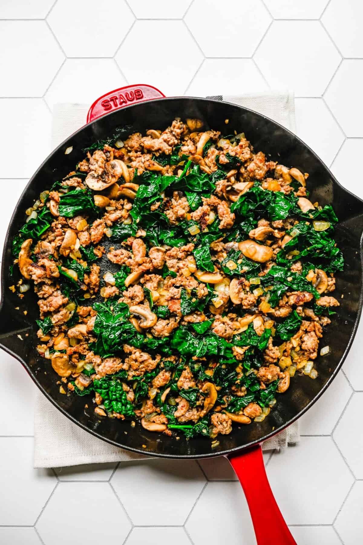A skillet filled with broken up Italian sausage, kale, mushrooms, onion, and garlic, cooked