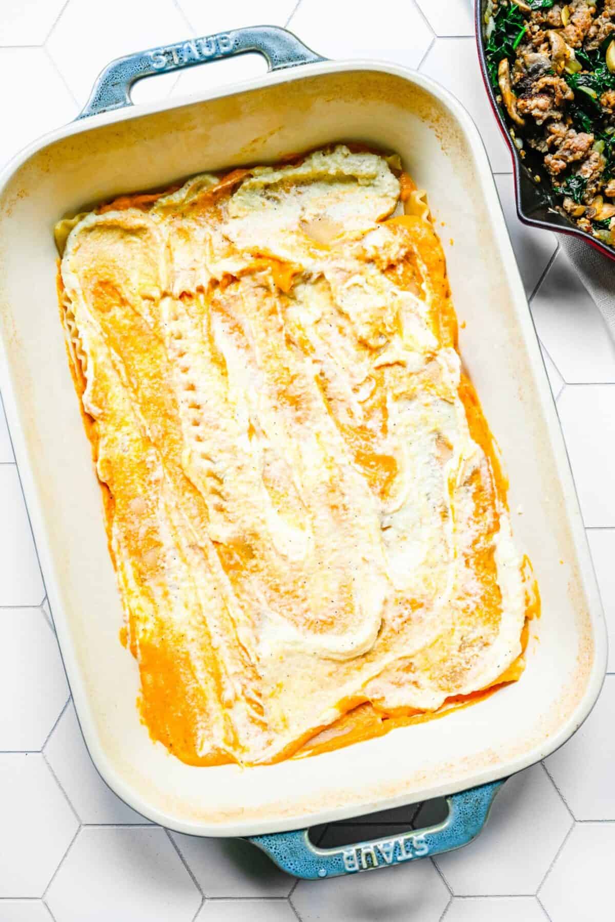 A baking dish with pumpkin cream sauce and ricotta filling spread over a layer of lasagna noodles