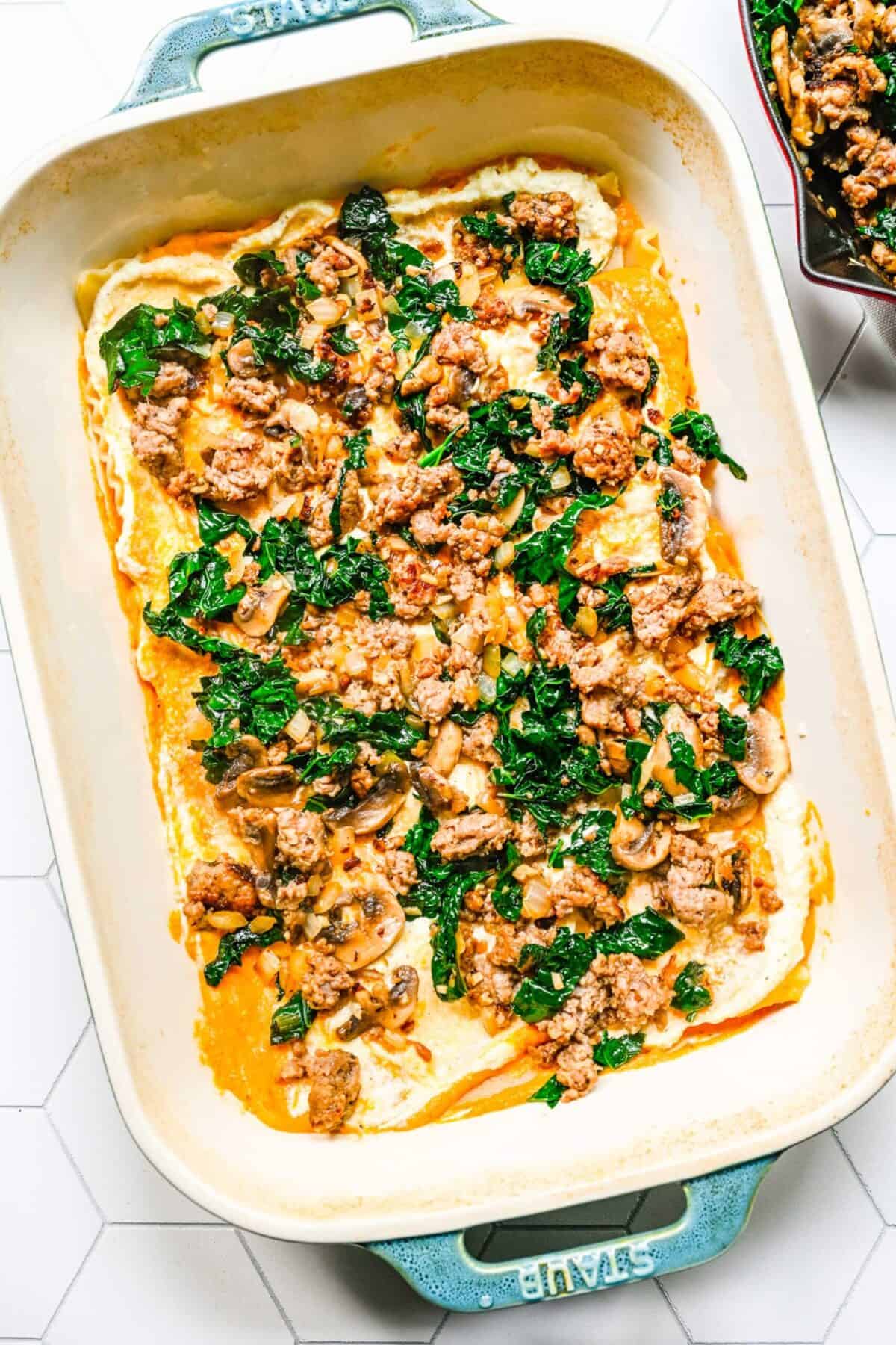 A baking dish with cooked sausage and kale spread over pumpkin cream sauce and ricotta filling, on top of lasagna noodles