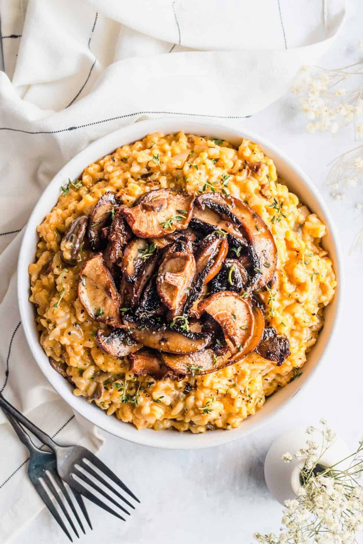 Overhead view of a serving bowl of risotto, topped with mushrooms, next to utensils, a kitchen towel, and a vase of flowers