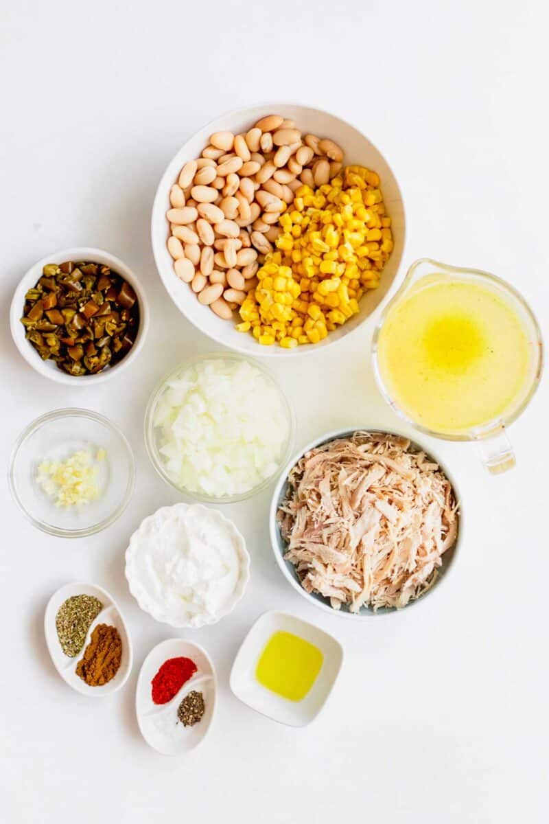Overhead shot of all ingredients for white turkey chili. Going clockwise: cannellini beans, corn, turkey stock, shredded turkey, olive oil, seasonings, cream cheese, diced onions, minced garlic, and chopped jalapenos