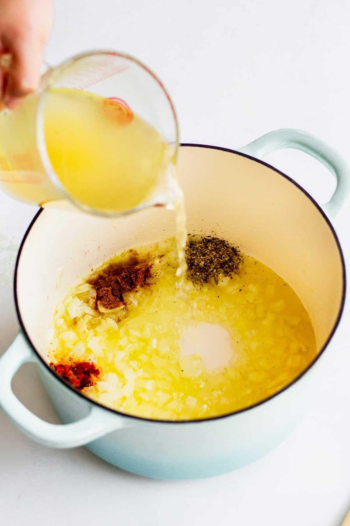 Pouring turkey stock into large pot with cooked onions and garlic. Seasonings such as oregano, cumin, and cayenne can be seen sprinkled into the soup.