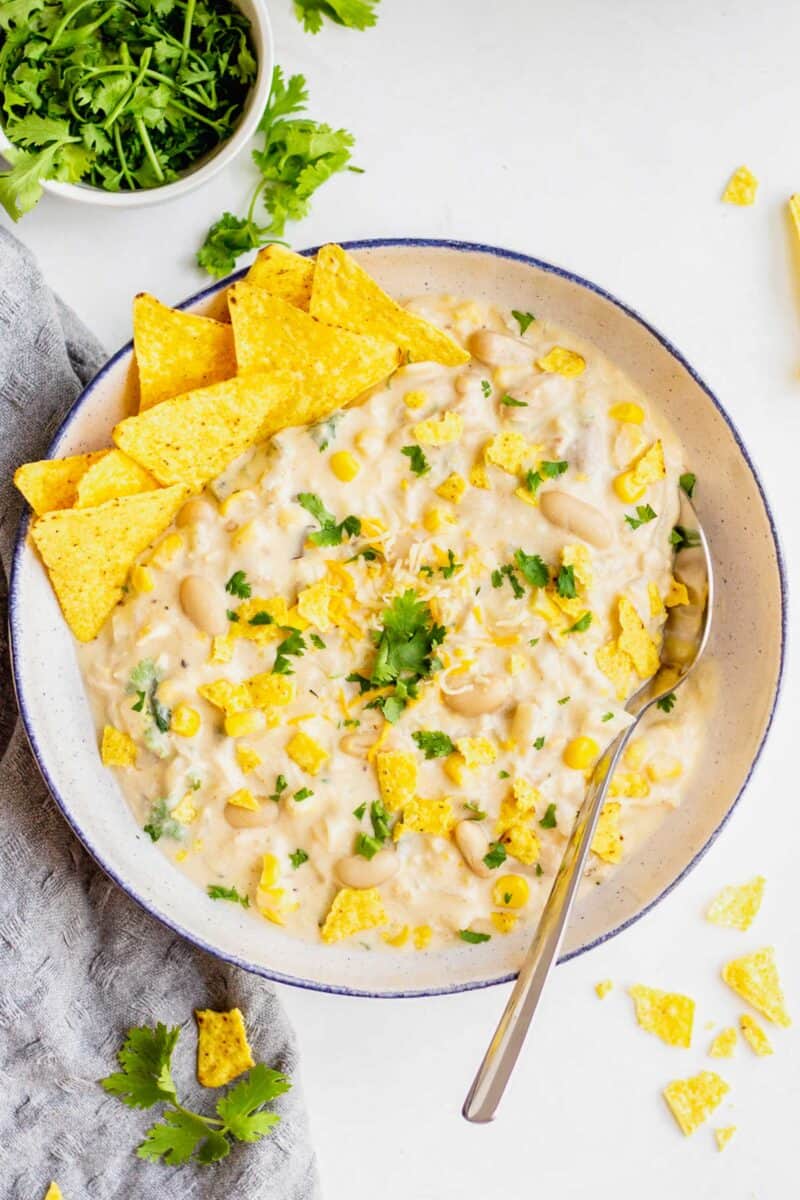 white turkey chili in a large white speckled bowl with a blue rim. cilantro can be seen above the bowl and some crushed up tortilla chips scattered around the bowl as well.