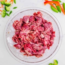 beef marinating in a clear bowl with cornstarch; broccoli, carrots, snow peas, and red bell pepper slices can be seen scattered around the marble