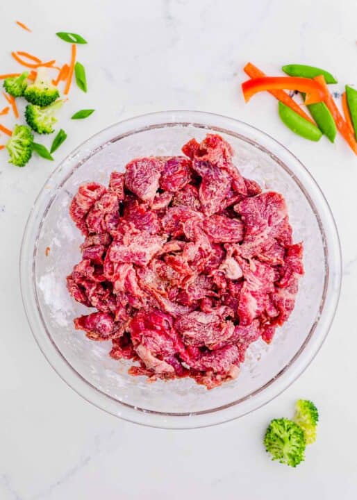 beef marinating in a clear bowl with cornstarch; broccoli, carrots, snow peas, and red bell pepper slices can be seen scattered around the marble