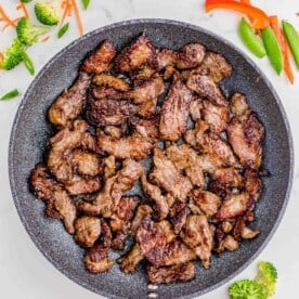 browned slices of beef in a grey nonstick skillet on a marble counter with broccoli, carrots, and other vegetables around