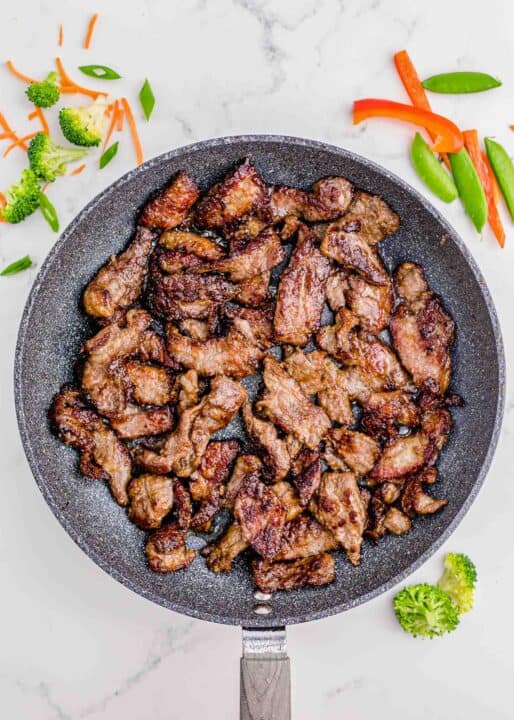 browned slices of beef in a grey nonstick skillet on a marble counter with broccoli, carrots, and other vegetables around