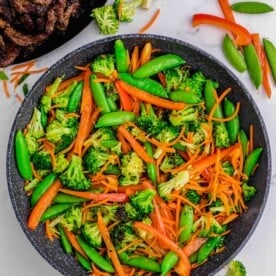 broccoli, sugar snap peas, carrots, and red bell peppers in a grey nonstick skillet on a marble countertop next to a plate of browned beef slices