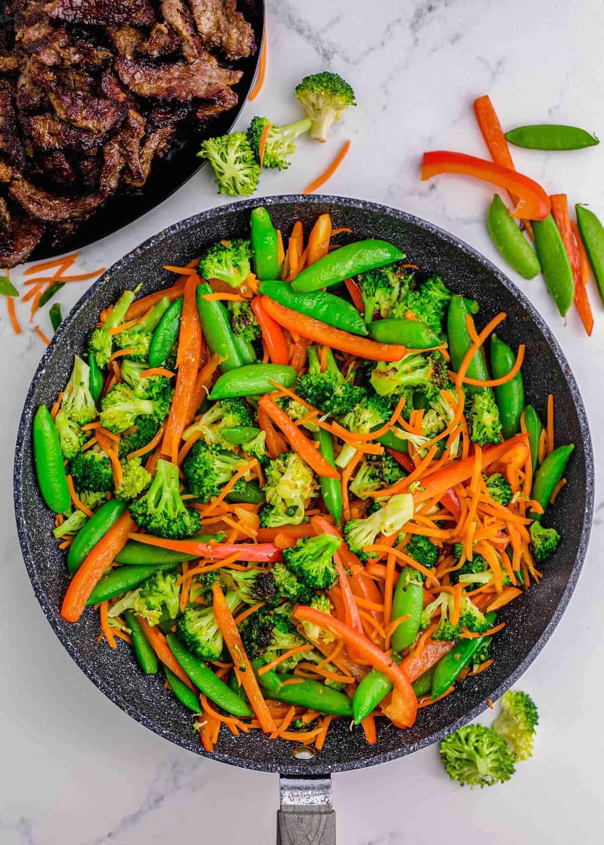 broccoli, sugar snap peas, carrots, and red bell peppers in a grey nonstick skillet on a marble countertop next to a plate of browned beef slices