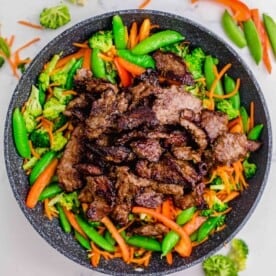 browned beef slices are added on top of cooked vegetables in a grey nonstick skillet