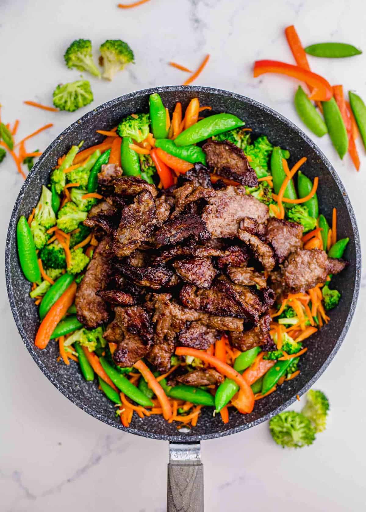 browned beef slices are added on top of cooked vegetables in a grey nonstick skillet