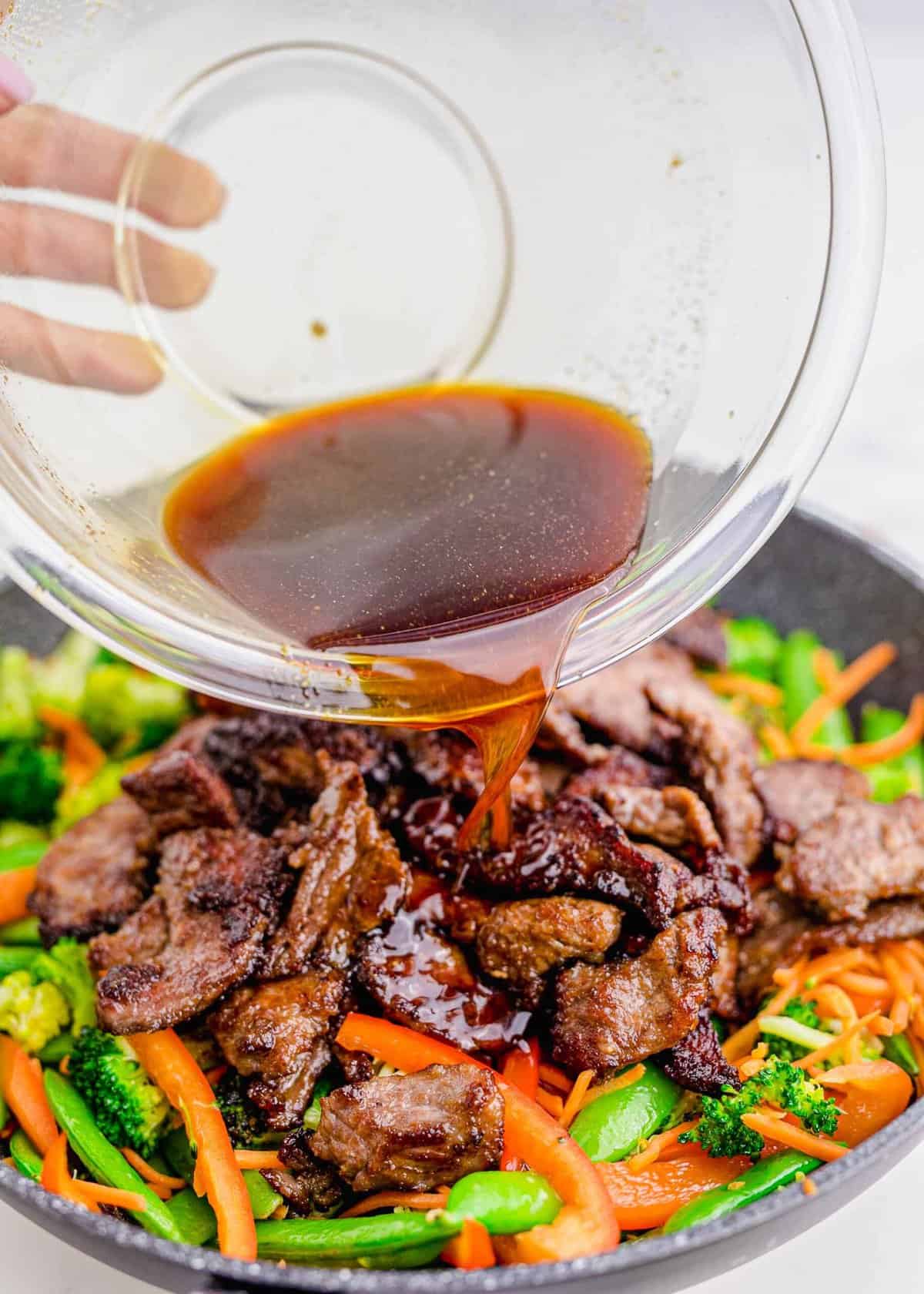 sauce is being poured on top of the browned beef slices and cooked vegetables in a grey nonstick skillet