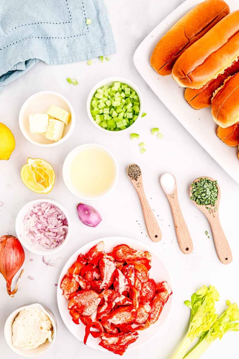 overhead image of lobster roll ingredients: celery, lemon juice, pepper, salt, minced chives, lobster meat, mayo, shallots, and butter. split top brioche buns can be seen on a white plate.