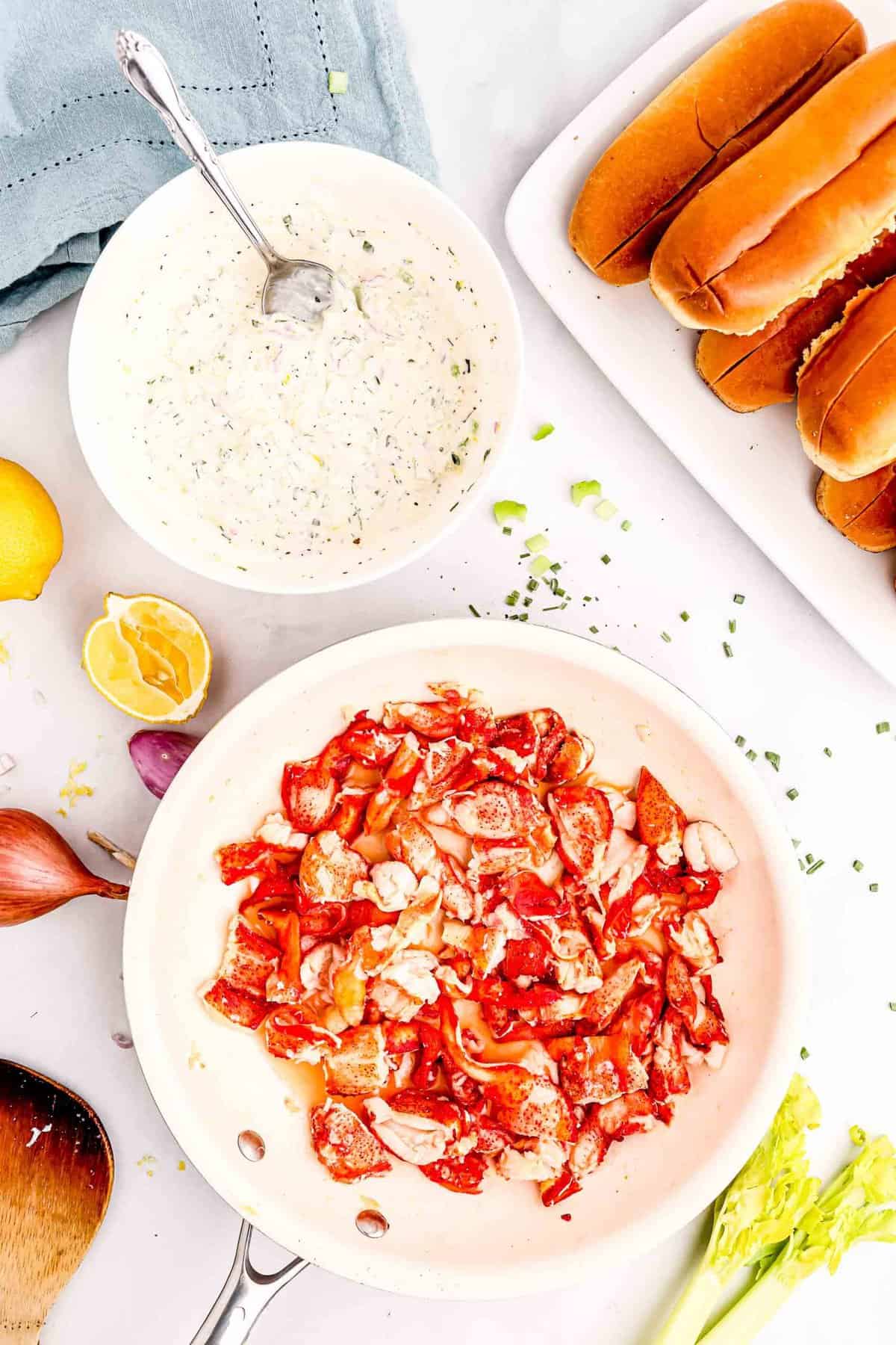 chunks of lobster meat in a skillet with butter next to a bowl of the mayo mixture and split top brioche buns