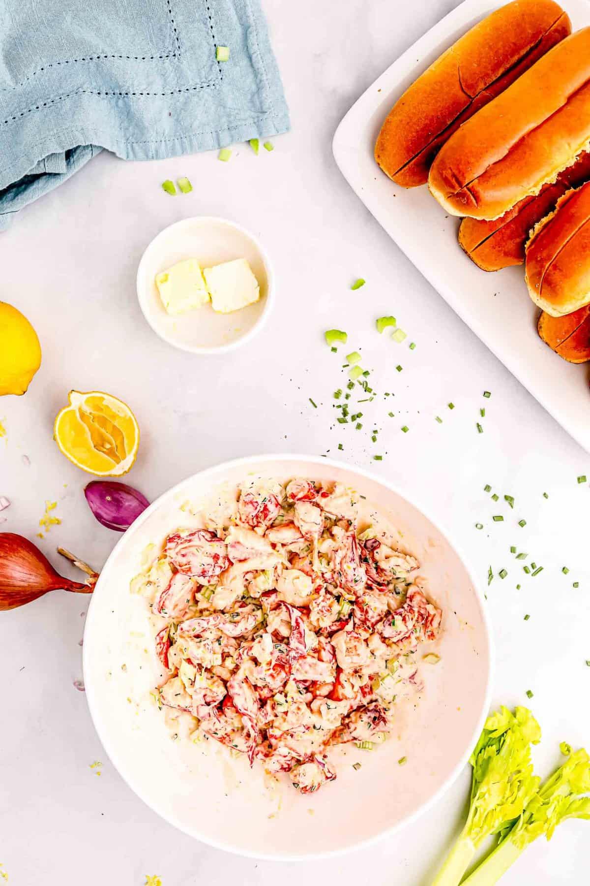 mayo mixture mixed in with the lobster meat in a white bowl. two pads of butter can be seen next to the bowl alongside the split top brioche buns