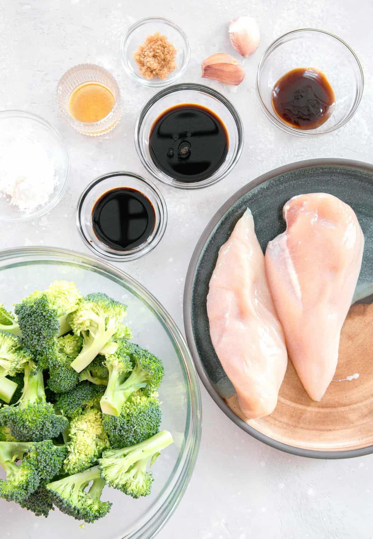 overhead image of ingredients for chinese chicken and broccoli: broccoli florets, dark soy sauce, cornstarch, sesame oil, brown sugar, low sodium soy sauce, garlic cloves, oyster sauce, and two boneless skinless chicken breasts