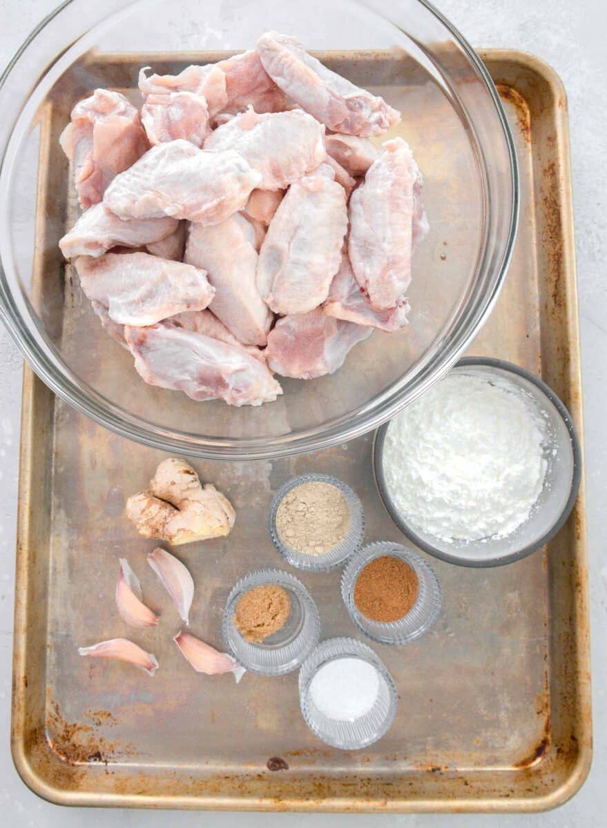 overhead image of a large baking sheet with ingredients for chinese salt and pepper chicken: chicken wings, cornstarch, chinese five spice powder, salt, brown sugar, white pepper powder, garlic, and ginger
