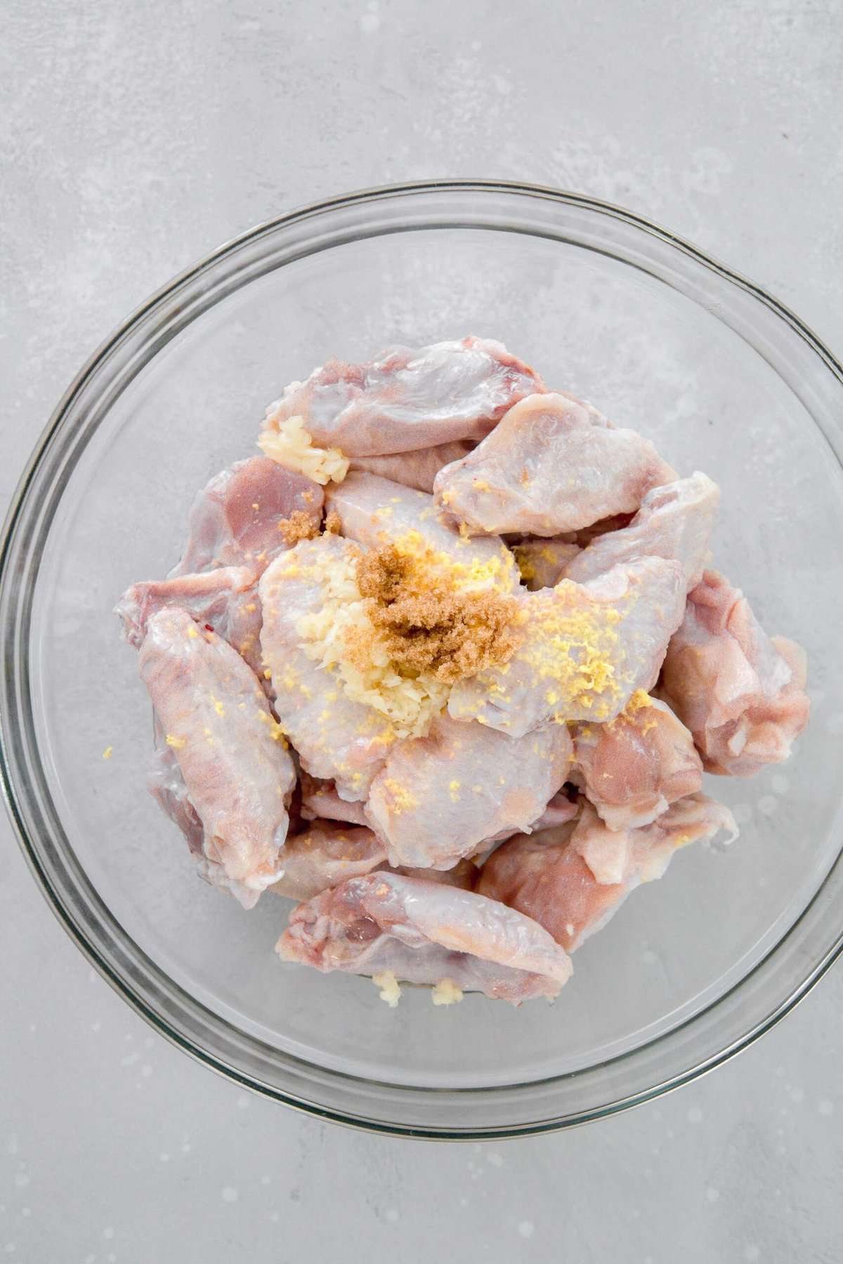 overhead image of chicken wings in a large clear bowl with garlic, ginger, and brown sugar on top