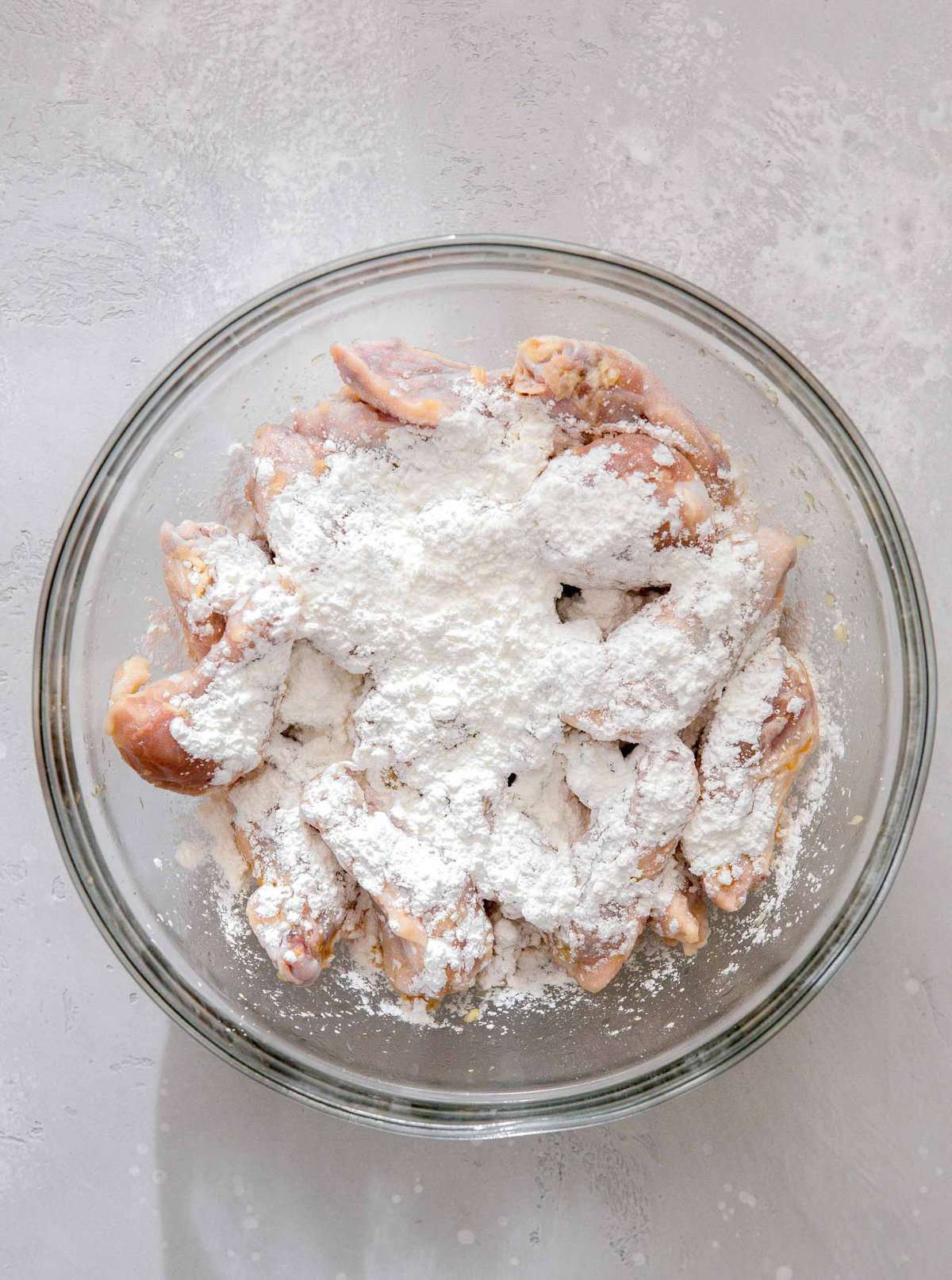 overhead image of chicken wings in a large clear bowl with cornstarch dumped on top of the wings