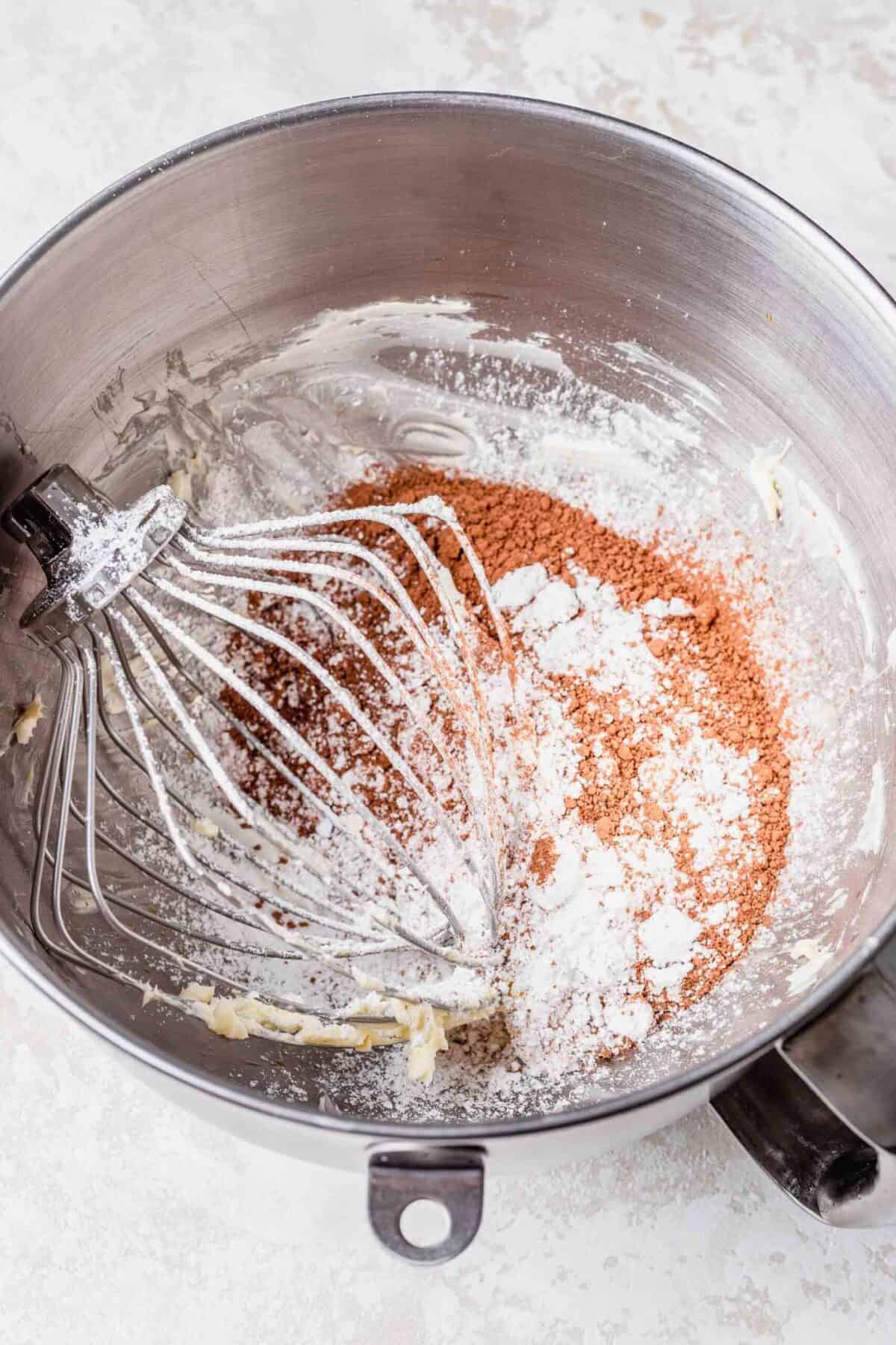 A stand mixer bowl filled with butter, sugar, cocoa powder, and cream, with a whisk attachment in the bowl