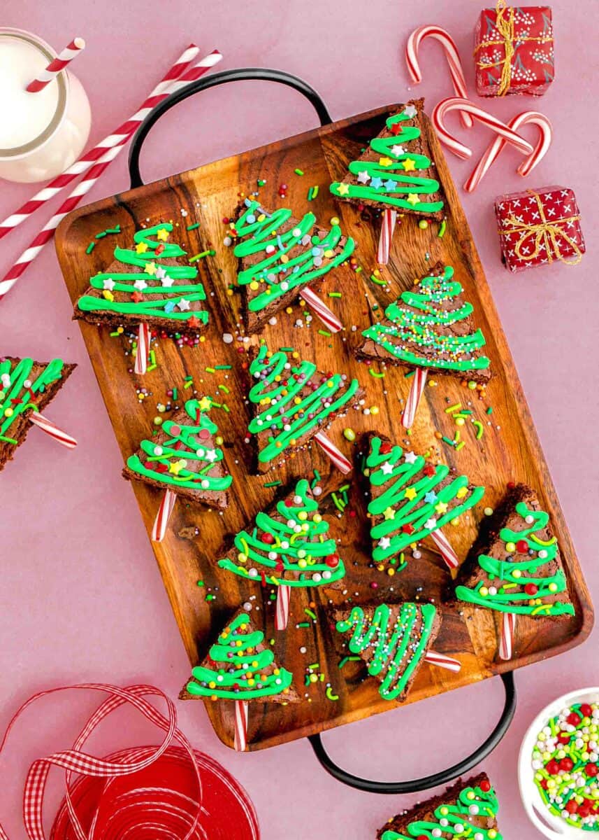 overhead image of christmas tree brownies on a wooden tray with faux presents next to it. ribbon, sprinkles, milk, and straws can be seen in the photo