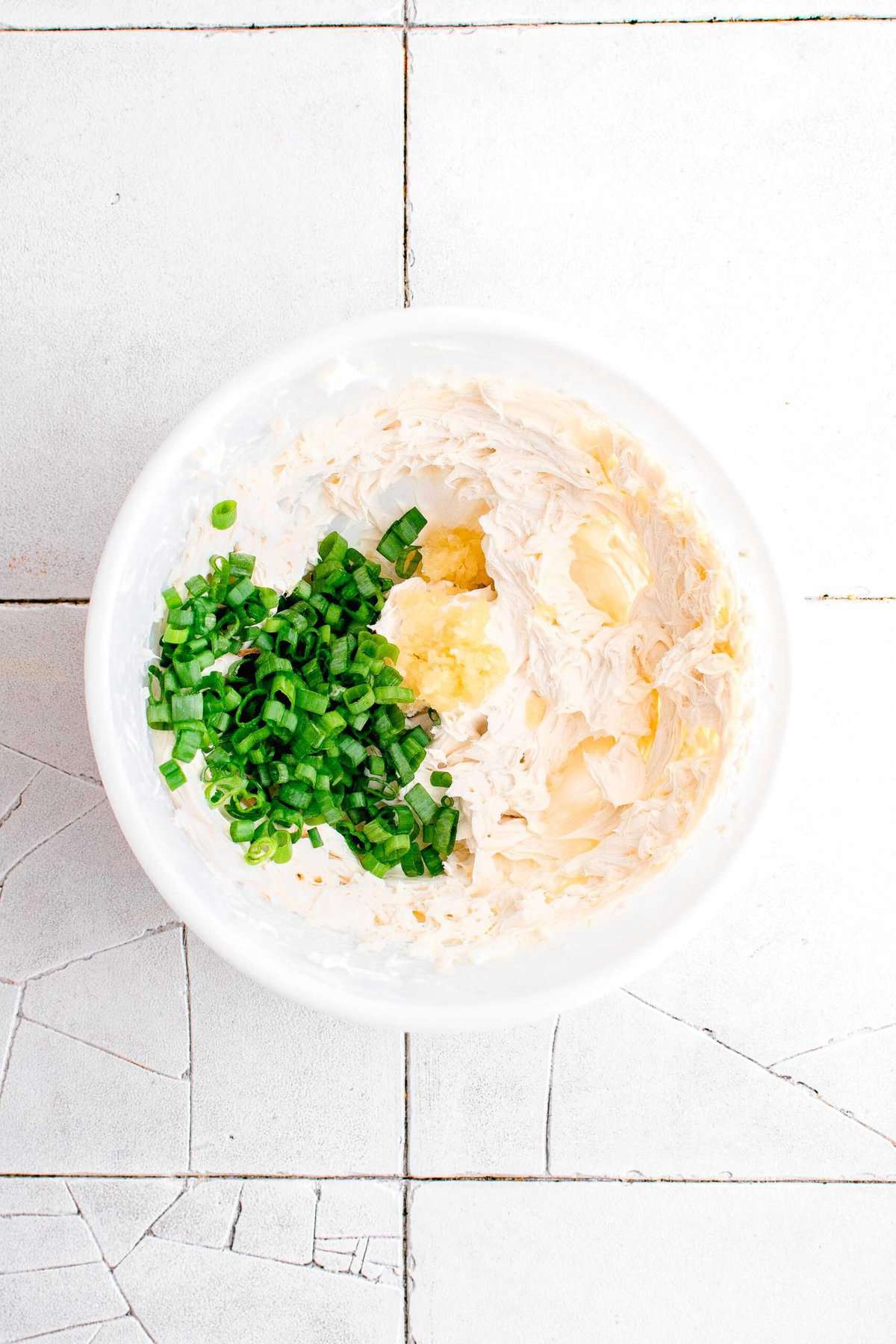 whipped cream cheese mixture in a white bowl on cracked grey tiles with scallions and garlic being added in