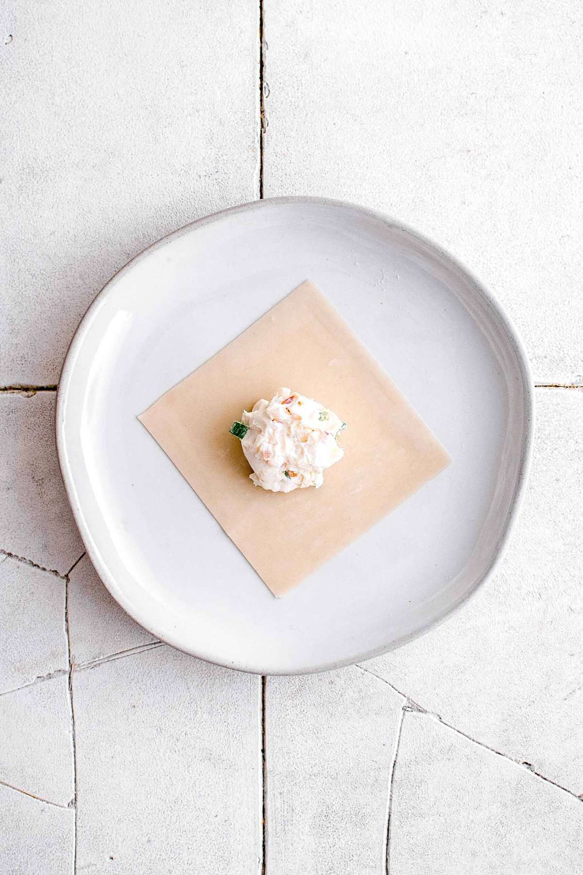 two teaspoons of crab rangoon filling in the center of a wonton wrapper on a white plate set on top of cracked grey tiles