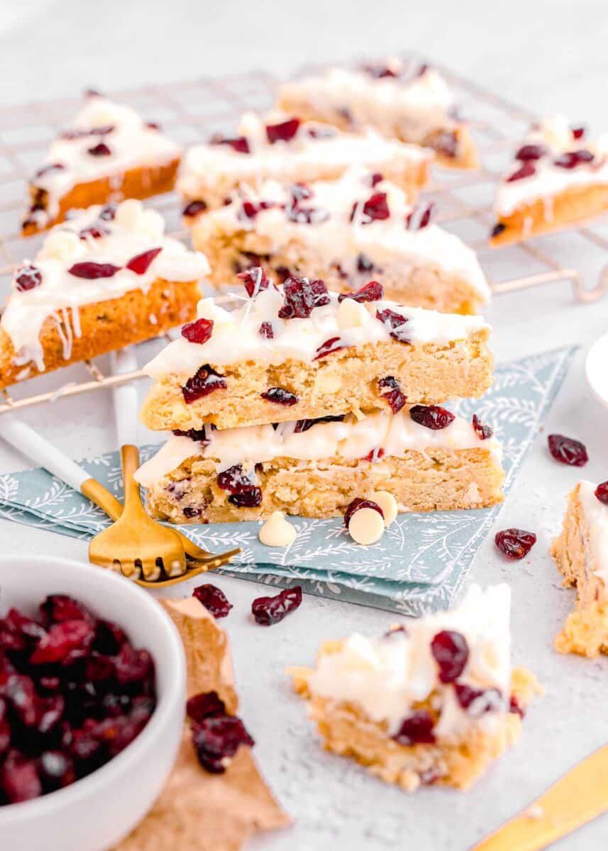 cranberry bliss bars stacked on top of a blue floral towel in front of a metal baking rack with more cranberry bliss bars on top