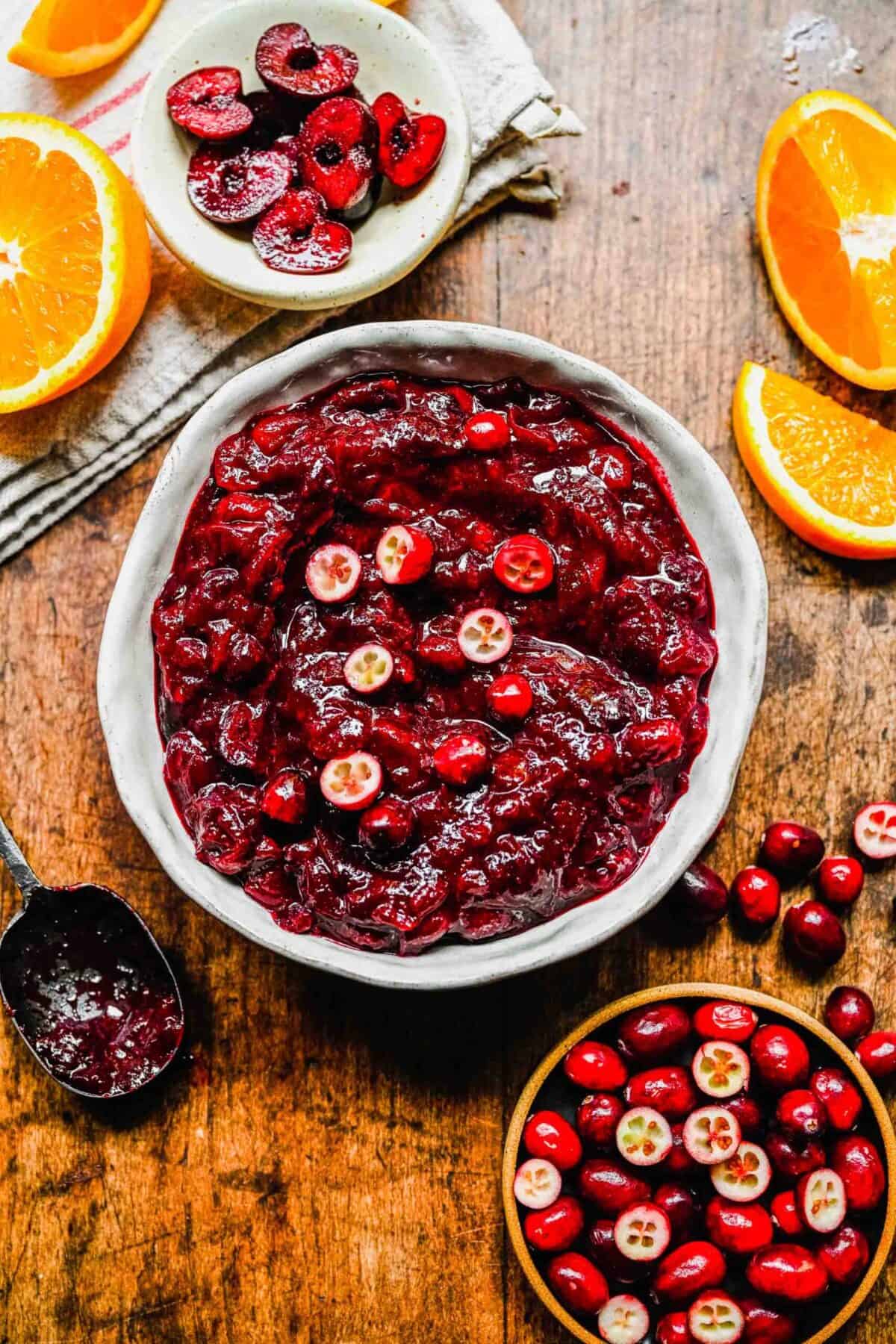 A bowl of cranberry cherry compote topped with fresh cranberries, next to a bowl of cranberries, a bowl of cherries, a dirty spoon, and some orange slices