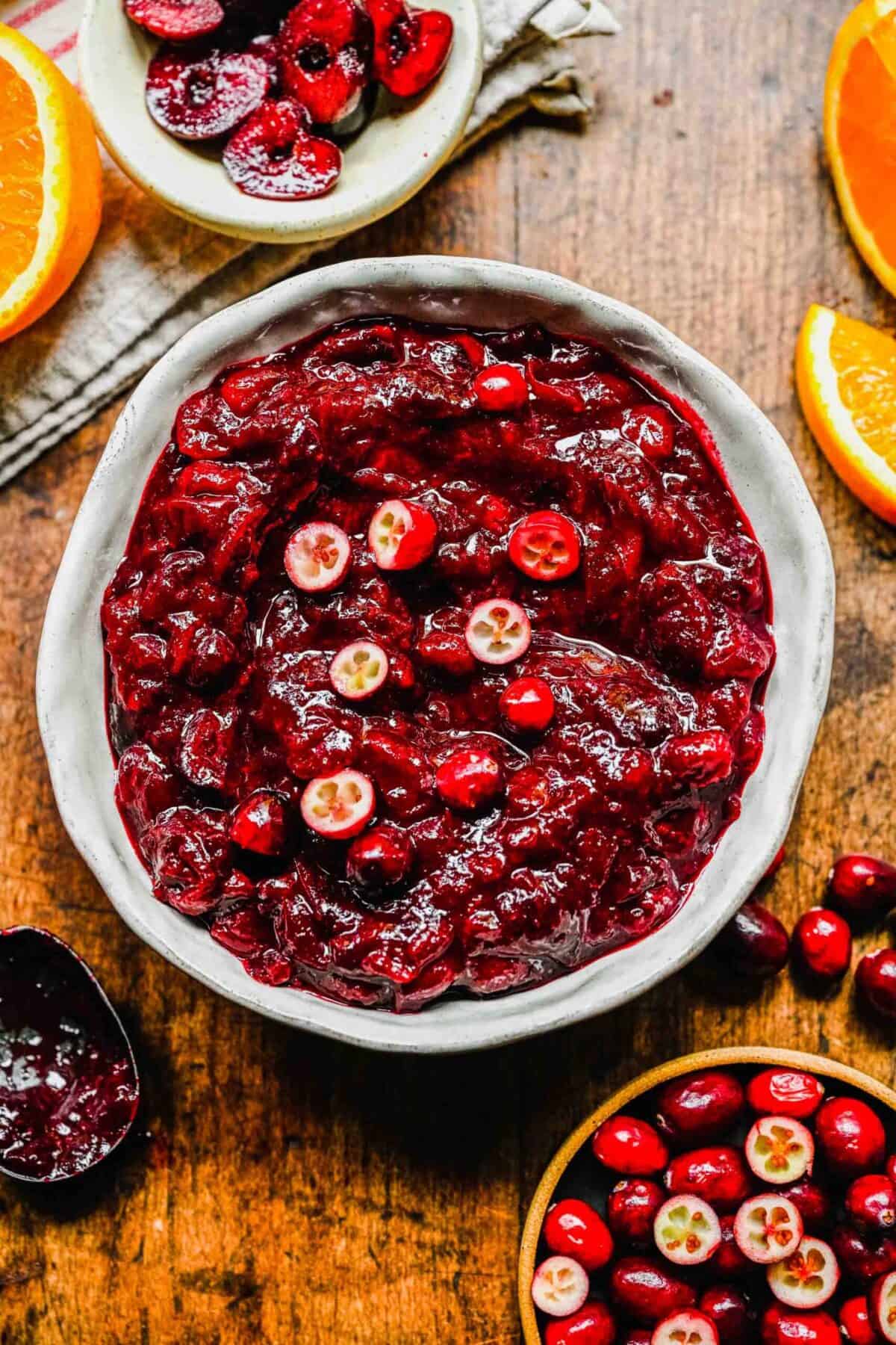 A bowl of cranberry cherry compote, topped with fresh cranberries, next to a bowl of cranberries and a bowl of cherries