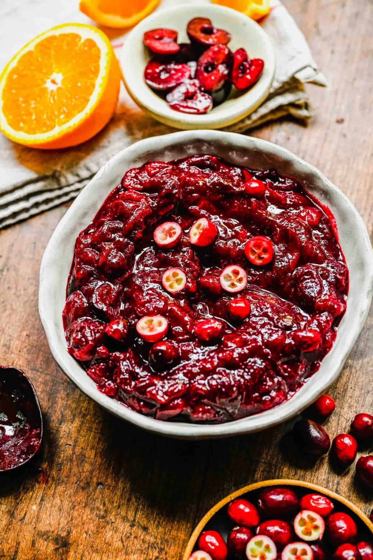 A bowl of cranberry cherry compote topped with sliced cranberries, next to half an orange, a bowl of cranberries, a bowl of cherries, and a dirty spoon