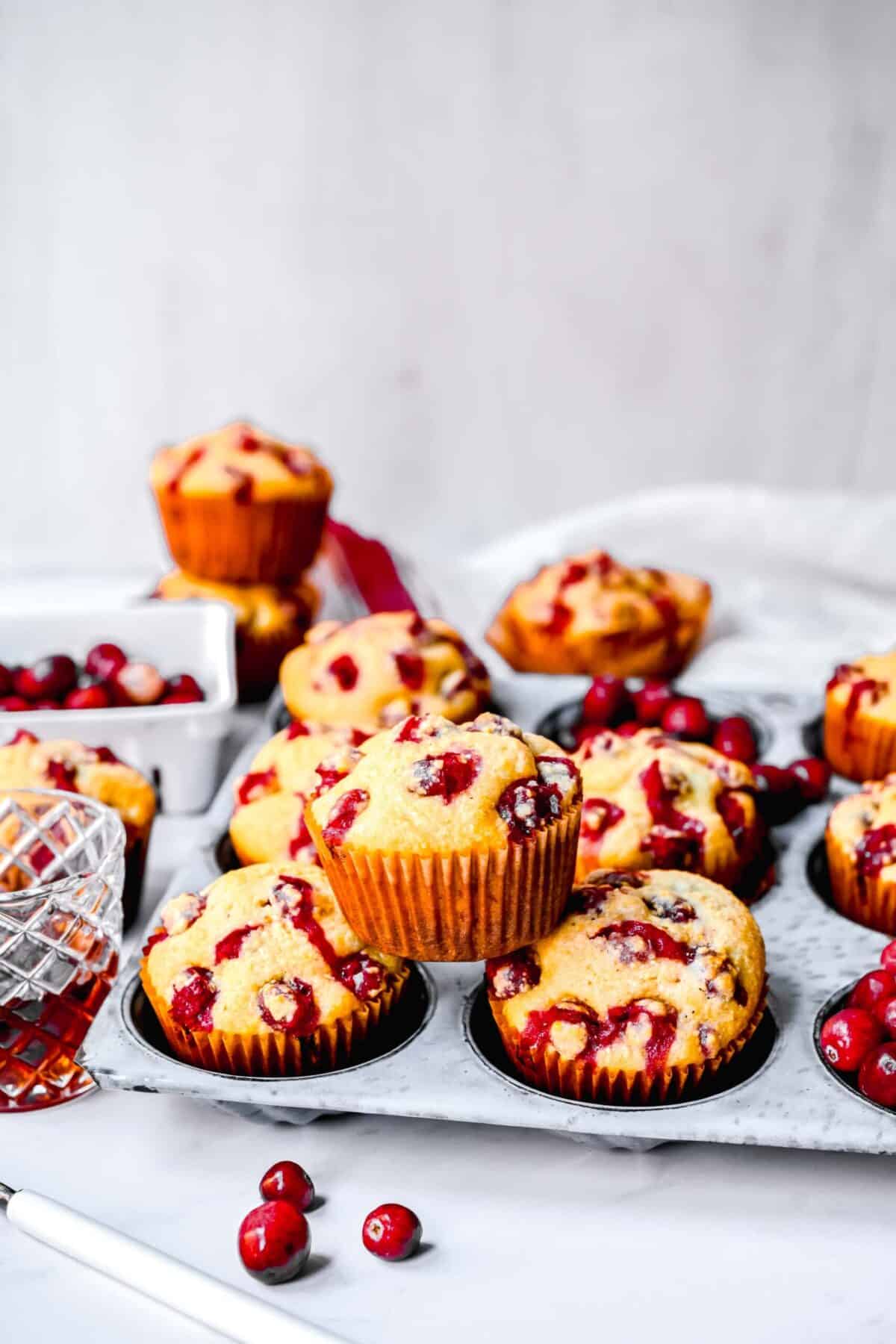 Cranberry cornbread muffins in a muffin tin, with a muffin resting on top of two of them, with muffins and cranberries in the background