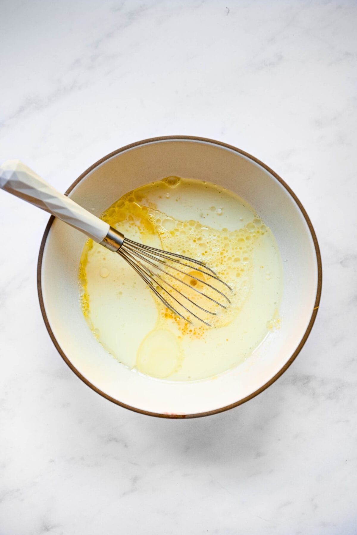 A mixing bowl with milk, oil, sugar, eggs, and a whisk in it