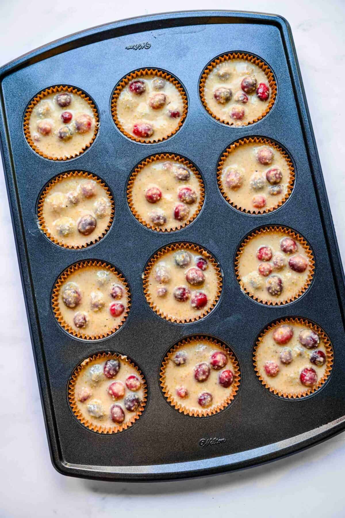 A muffin tin with 12 uncooked cranberry cornbread muffins in it