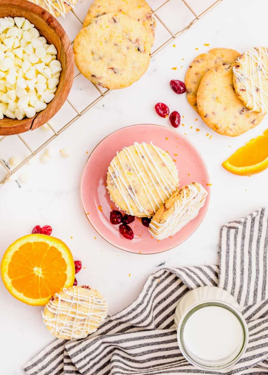 overhead image of cranberry shortbread cookie on a pink plate next to an orange, bowl of white chocolate chips, and more cookies. a towel with a glass of milk and linen striped towel can be seen as well