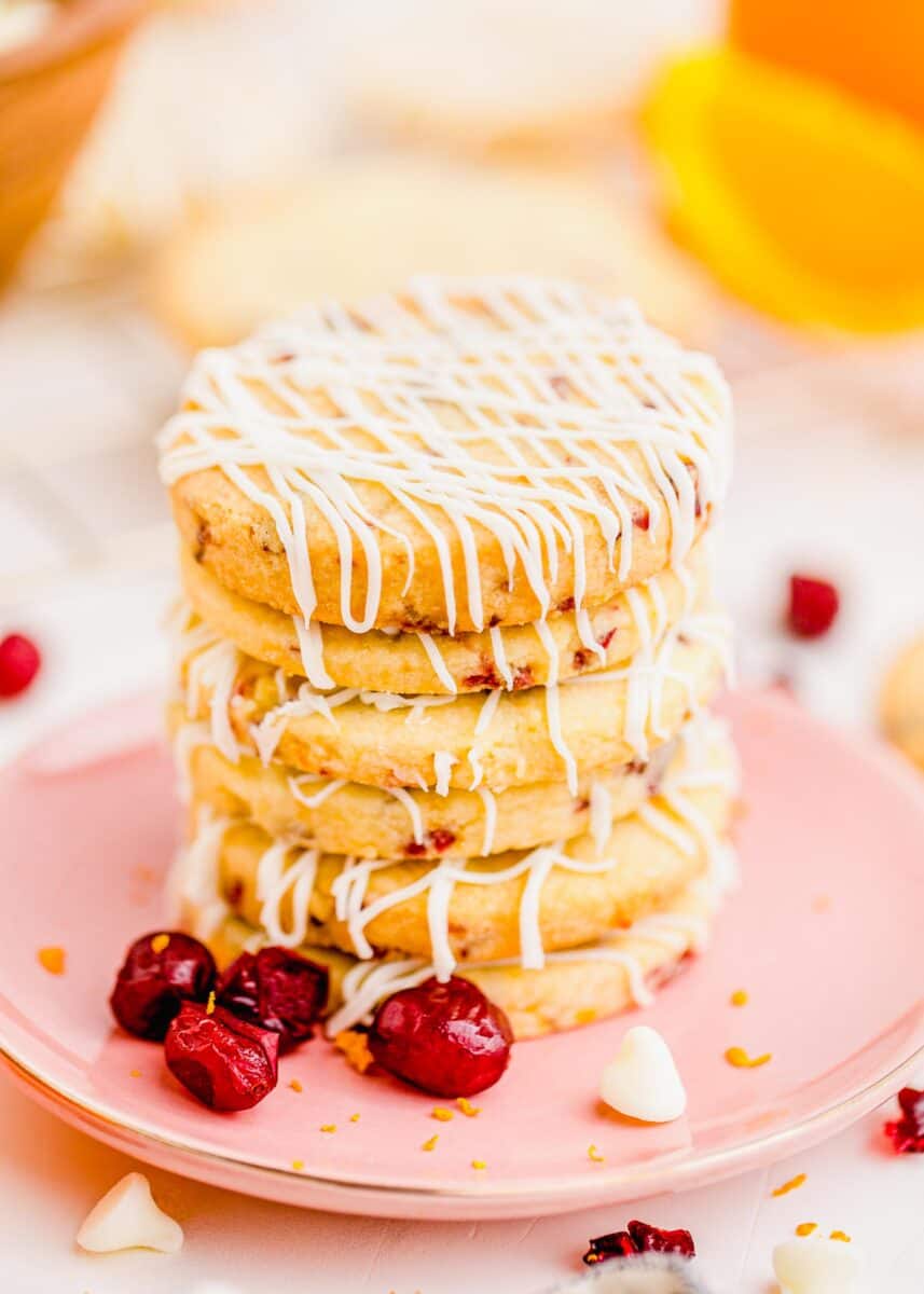 stacked cranberry orange shortbread cookies with white chocolate drizzle stacked on a pink plate with dried cranberries and white chocolate