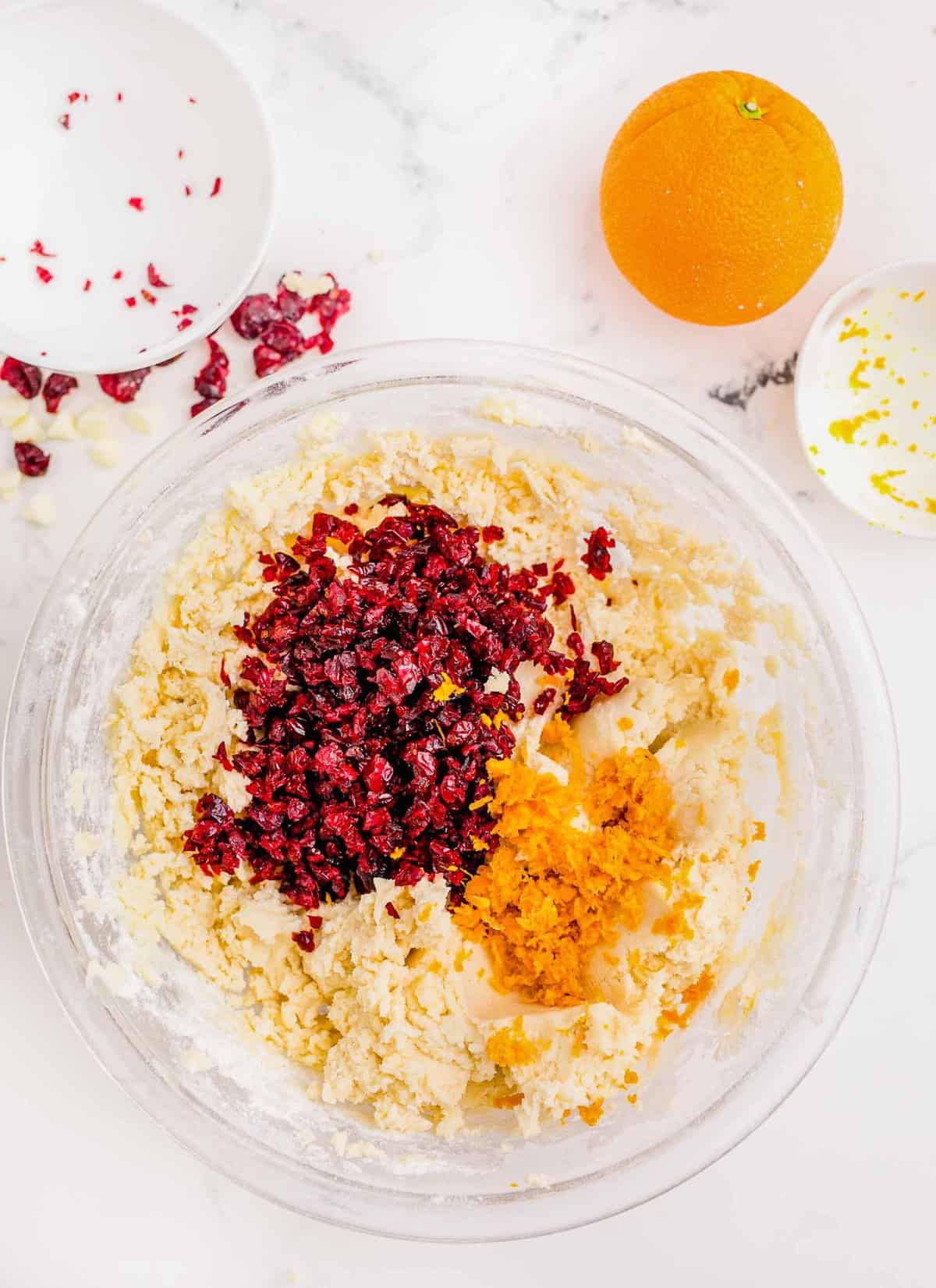dried cranberries and orange zest is added to shortbread cookie dough in a large clear bowl