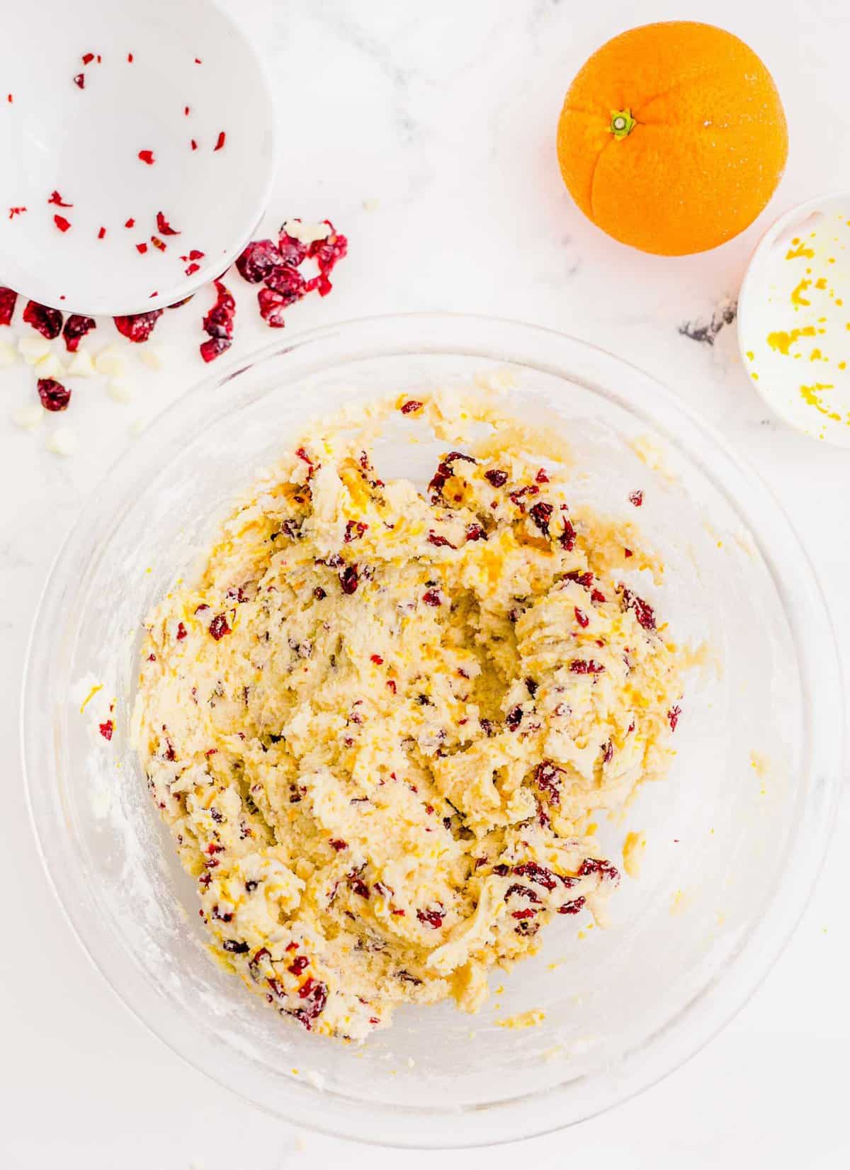 cranberry orange shortbread cookie dough mixed with dried cranberries and orange zest in a large clear bowl next to an orange