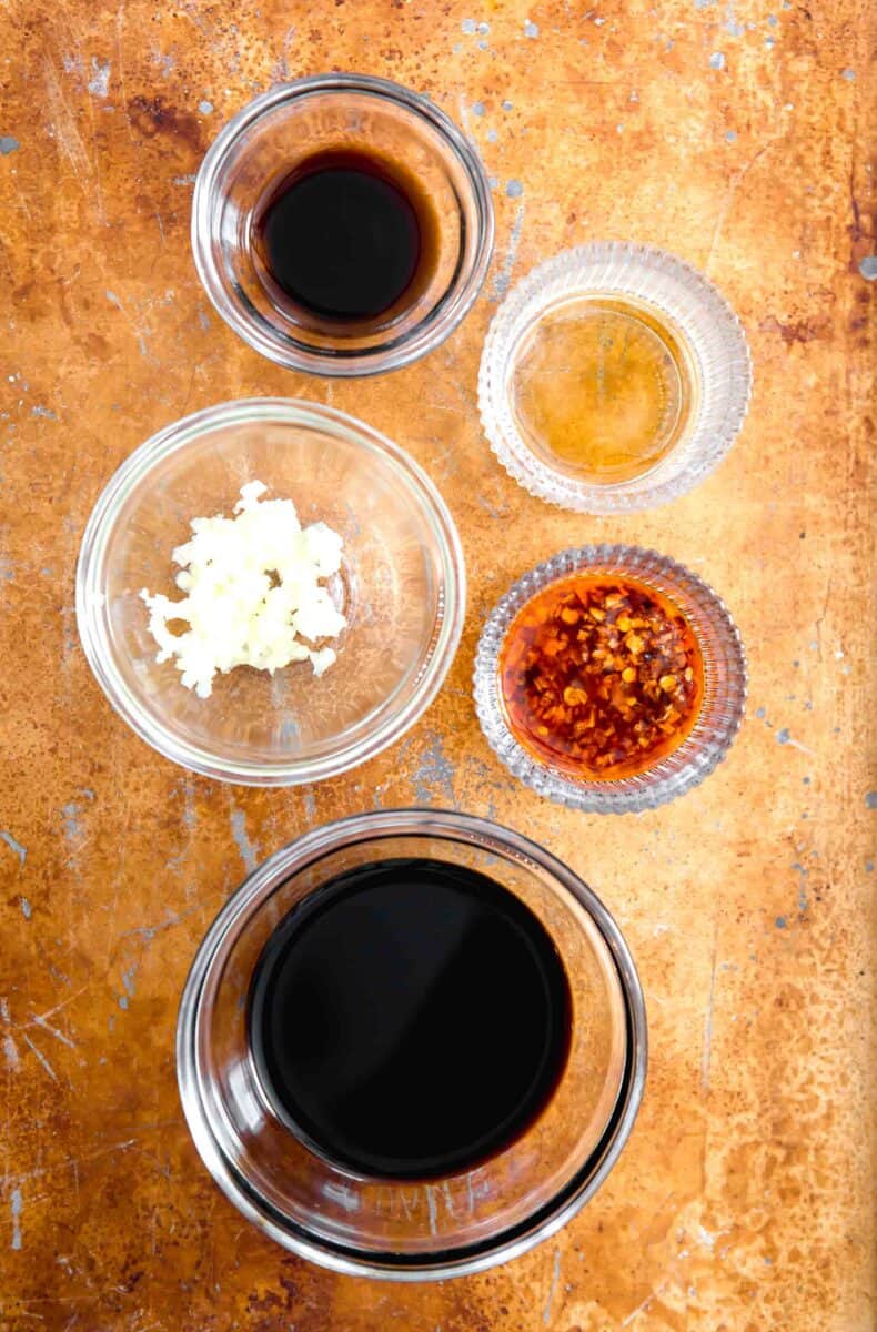 overhead image of ingredients for dumpling sauce: black vinegar, sesame oil, chili crisp, sweet soy sauce, and fresh minced garlic