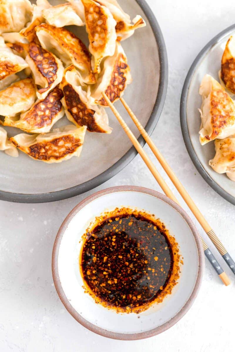 overhead image of dumpling sauce in a brown rimmed white ceramic bowl next to two grey plates of pan-fried dumplings. wooden chopsticks can be seen resting on the grey plate.