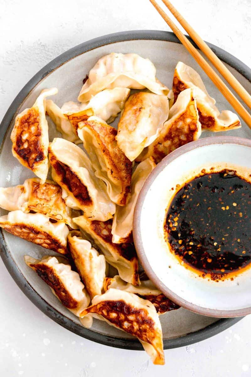 overhead image of pan-fried dumplings on a grey ceramic plate with the dumpling sauce on the plate next to wooden chopsticks
