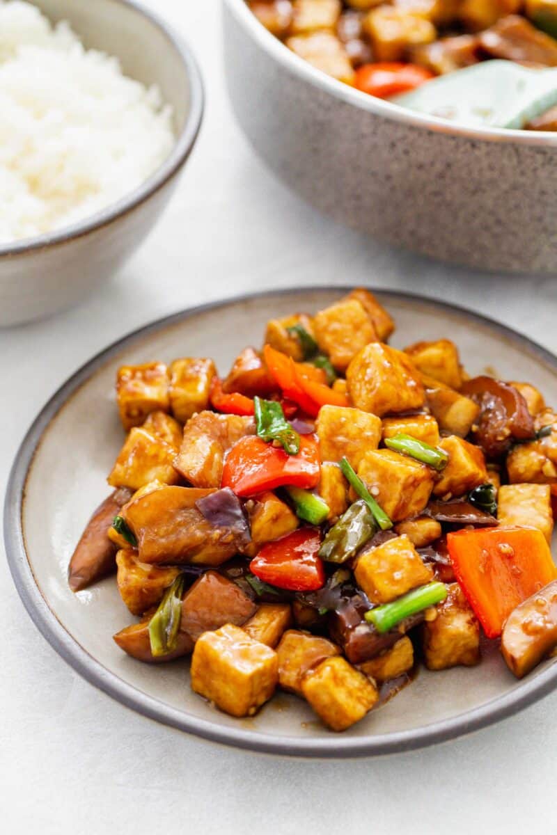 eggplant tofu dish on a grey ceramic plate with dark grey rim. a bowl of rice and the skillet with more tofu eggplant can be seen in the background